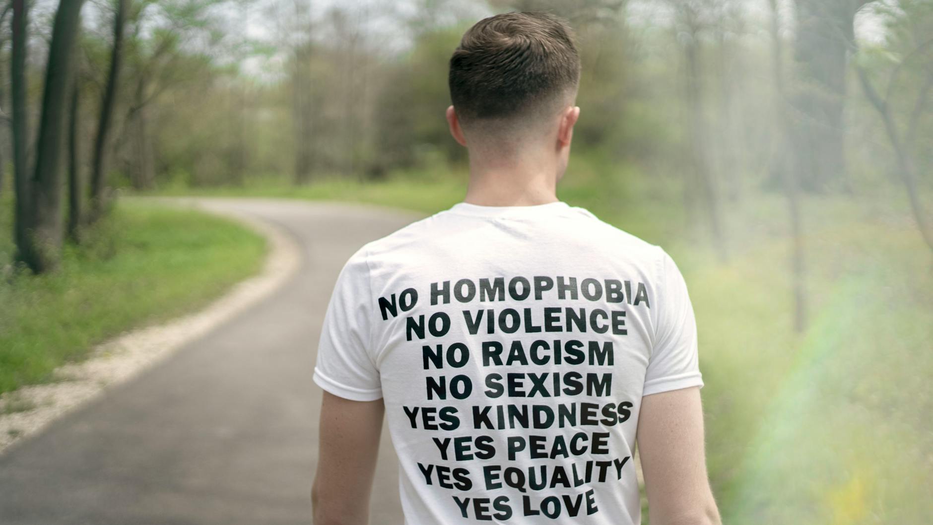 A person walking away from the camera in a white t-shirt with the words no homophobia, violence, racism, sexism and yes kindness, peace, equality, love