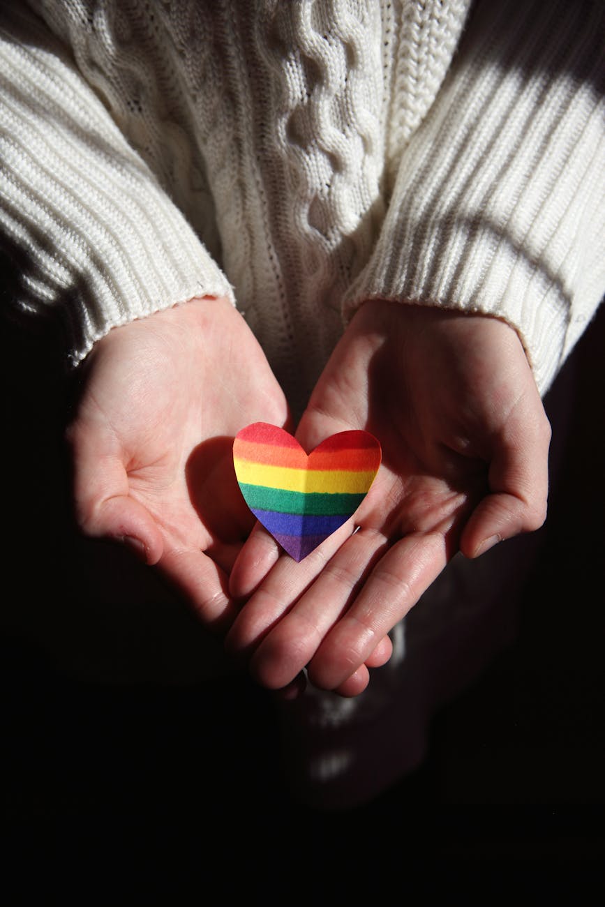 A pair of hands holding a paper heart that is rainbow colors